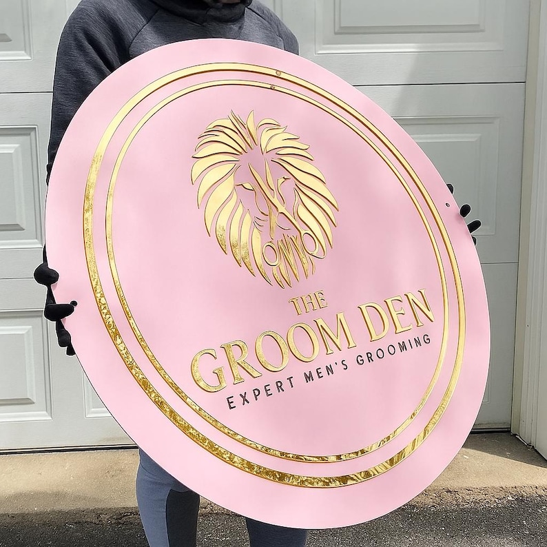 May include: Pink, oval sign with gold trim and a gold lion emblem. The sign reads "THE GROOM DEN EXPERT MEN'S GROOMING" in gold lettering. The sign is held up by a person.