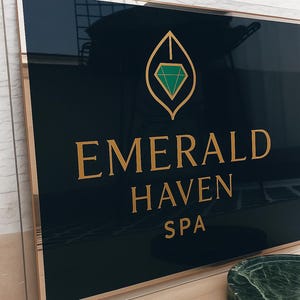 May include: A rectangular sign with a black background and gold trim. The sign reads "EMERALD HAVEN SPA" in gold lettering. A gold leaf design with a green diamond is at the top. The sign is in a clear acrylic frame.