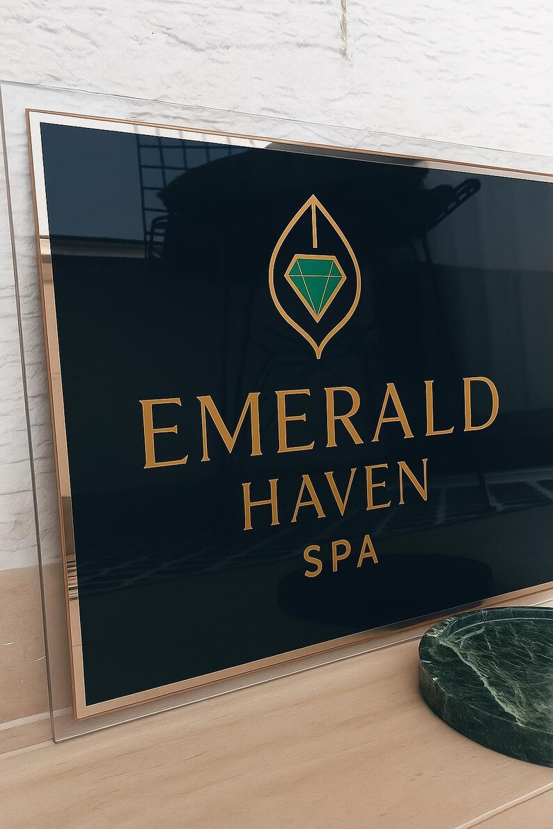 May include: A rectangular sign with a black background and gold trim. The sign reads "EMERALD HAVEN SPA" in gold lettering. A gold leaf design with a green diamond is at the top. The sign is in a clear acrylic frame.