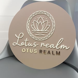 May include: A circular sign featuring the words "Lotus realm" in a cursive font and "OTUS REALM" in gold block letters. The sign has a beige background and a white lotus flower design.
