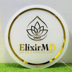 K&ouml;nnte beinhalten: Ein rundes, wei&szlig;es Schild mit goldenem Rand und goldener Schrift. Das Schild zeigt den Text "Elixir MD" und ein Lotusblumen-Logo. Der Text "MED SPA & HYDRATION CLINIC" ist ebenfalls enthalten.