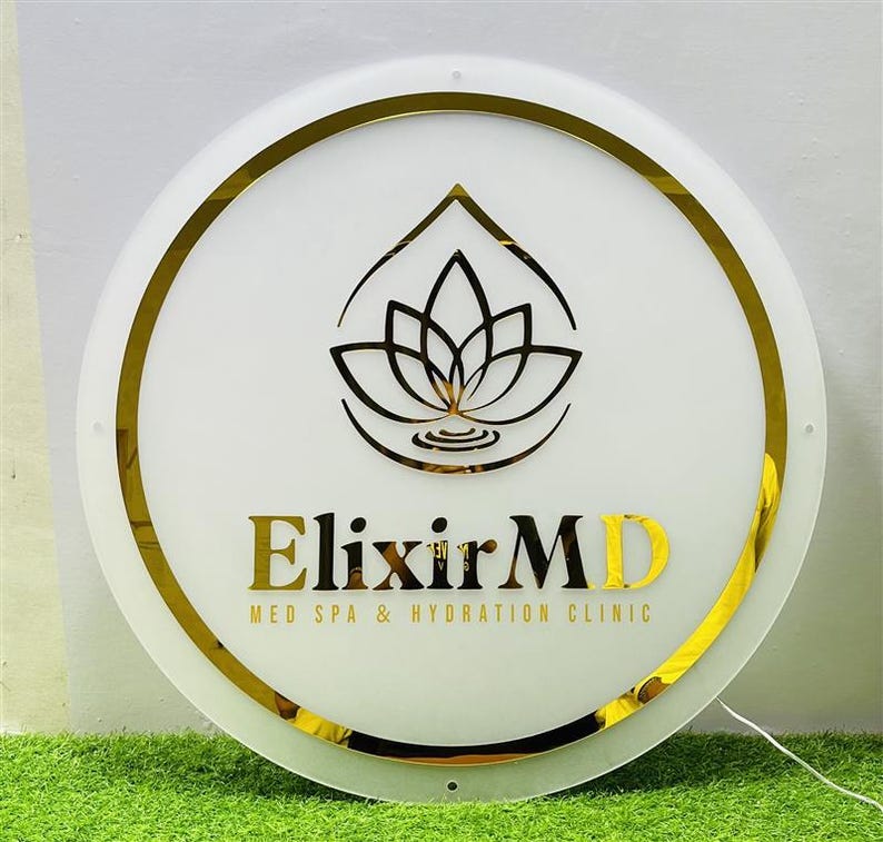 K&ouml;nnte beinhalten: Ein rundes, wei&szlig;es Schild mit goldenem Rand und goldener Schrift. Das Schild zeigt den Text "Elixir MD" und ein Lotusblumen-Logo. Der Text "MED SPA & HYDRATION CLINIC" ist ebenfalls enthalten.