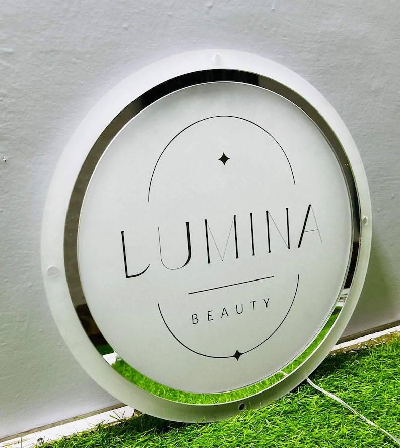 K&ouml;nnte beinhalten: Ein rundes, beleuchtetes Schild mit dem Text "LUMINA BEAUTY" in Schwarz. Das Schild hat einen wei&szlig;en Rahmen mit einem silbernen Innenring. Der Hintergrund ist eine hellgr&uuml;ne Oberfl&auml;che.