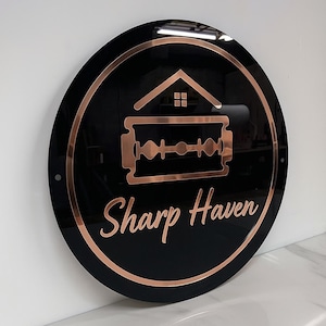 May include: A circular black sign with a copper-coloured border and logo. The logo shows a house outline above a razor blade, with the words "Sharp Haven" in a cursive font. The sign is mounted on a white wall.