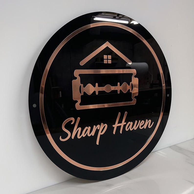 May include: A circular black sign with a copper-coloured border and logo. The logo shows a house outline above a razor blade, with the words "Sharp Haven" in a cursive font. The sign is mounted on a white wall.