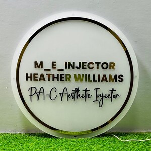 K&ouml;nnte beinhalten: Ein runder, wei&szlig;er Leuchtschild mit goldenem Rand. Der Schriftzug "M_E_INJECTOR", "HEATHER WILLIAMS" und "PA-C Aesthetic Injector" ist in verschiedenen Schriftarten und Farben dargestellt. Der Hintergrund ist gr&uuml;n.