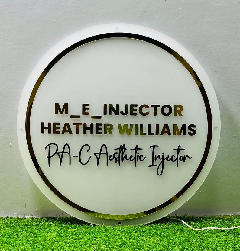 K&ouml;nnte beinhalten: Ein runder, wei&szlig;er Leuchtschild mit goldenem Rand. Der Schriftzug "M_E_INJECTOR", "HEATHER WILLIAMS" und "PA-C Aesthetic Injector" ist in verschiedenen Schriftarten und Farben dargestellt. Der Hintergrund ist gr&uuml;n.