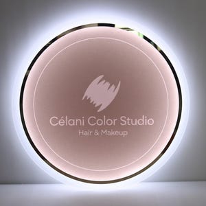 May include: A round, illuminated sign with a soft pink center and a white outer ring. The sign features the text "C&eacute;lani Color Studio" and "Hair & Makeup" in a clean, modern font, along with a stylized logo.