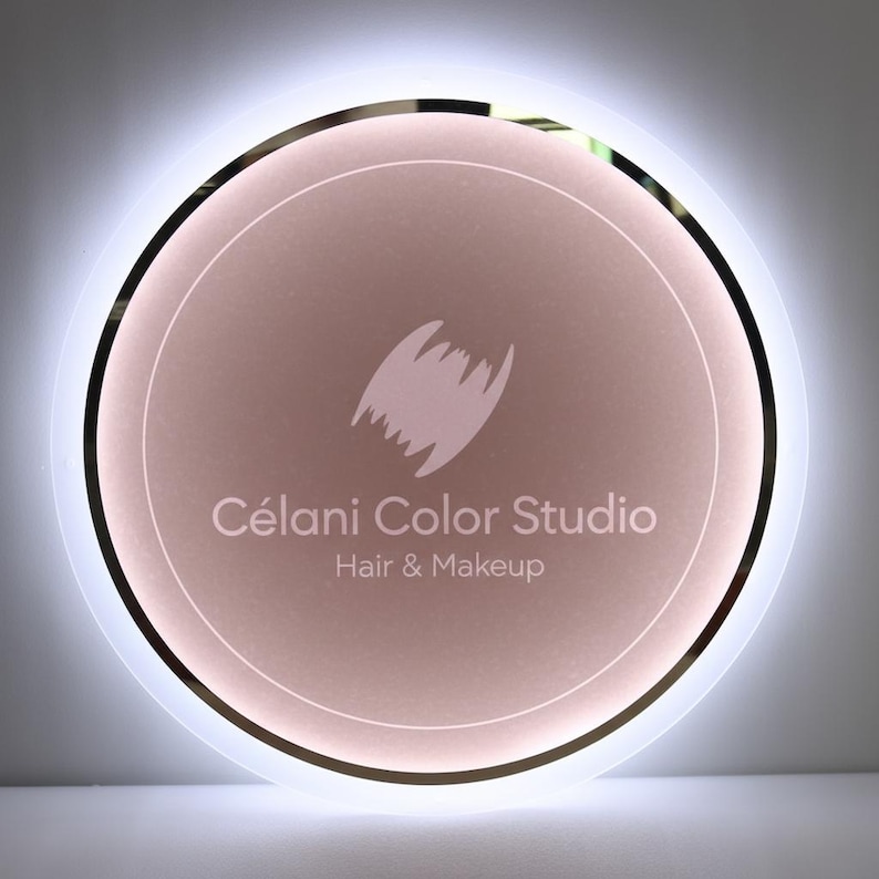 May include: A round, illuminated sign with a soft pink center and a white outer ring. The sign features the text "C&eacute;lani Color Studio" and "Hair & Makeup" in a clean, modern font, along with a stylized logo.