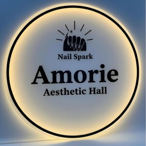 May include: A circular illuminated sign with a black border. The sign features the text "Nail Spark" above the word "Amorie" and "Aesthetic Hall" in a bold, black font. A nail design is at the top.