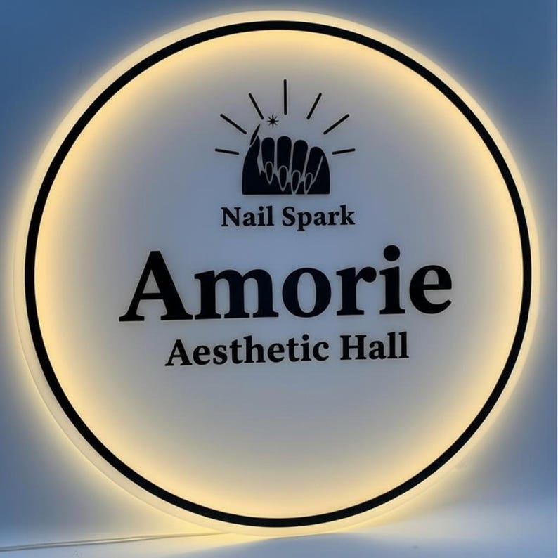 May include: A circular illuminated sign with a black border. The sign features the text "Nail Spark" above the word "Amorie" and "Aesthetic Hall" in a bold, black font. A nail design is at the top.