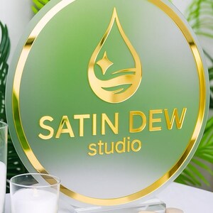 May include: A round sign with a gold border and the words "SATIN DEW studio" in gold lettering. The sign has a green background with a gold water droplet design. The sign is displayed on a clear stand with small stones in front of it.