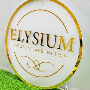 May include: A circular sign with a white face and gold trim. The sign displays the gold text "ELYSIUM MEDICAL AESTHETICS" and decorative gold lines. The sign is set against a backdrop of green grass.