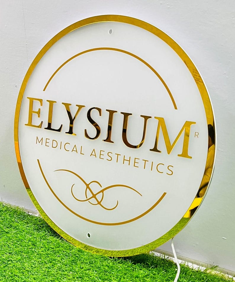 May include: A circular sign with a white face and gold trim. The sign displays the gold text "ELYSIUM MEDICAL AESTHETICS" and decorative gold lines. The sign is set against a backdrop of green grass.