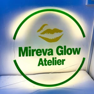 May include: A round, illuminated sign with a green border and the words "Mireva Glow Atelier" in green. The sign features a yellow lip graphic. The background is a soft, glowing white.