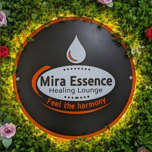 May include: A round black sign with the words "Mira Essence Healing Lounge" in white and orange. The sign has a white teardrop shape at the top and the words "Feel the harmony" at the bottom. The sign is surrounded by green foliage and flowers.