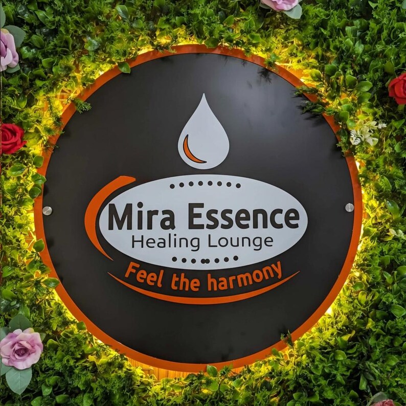 May include: A round black sign with the words "Mira Essence Healing Lounge" in white and orange. The sign has a white teardrop shape at the top and the words "Feel the harmony" at the bottom. The sign is surrounded by green foliage and flowers.