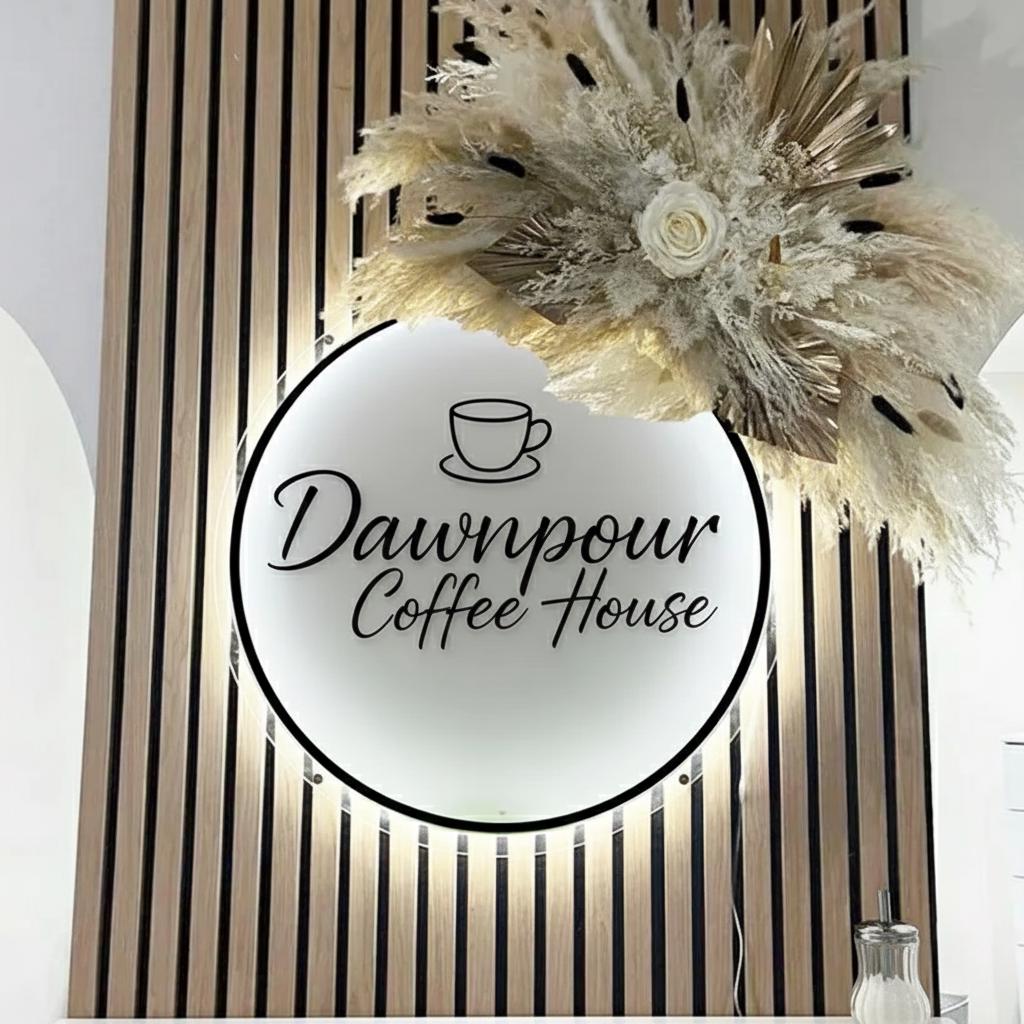 May include: A round, illuminated sign for "Dawnpour Coffee House" with a black border and the shop's name in elegant script. The sign is set against a wooden slatted wall and adorned with dried floral arrangements.