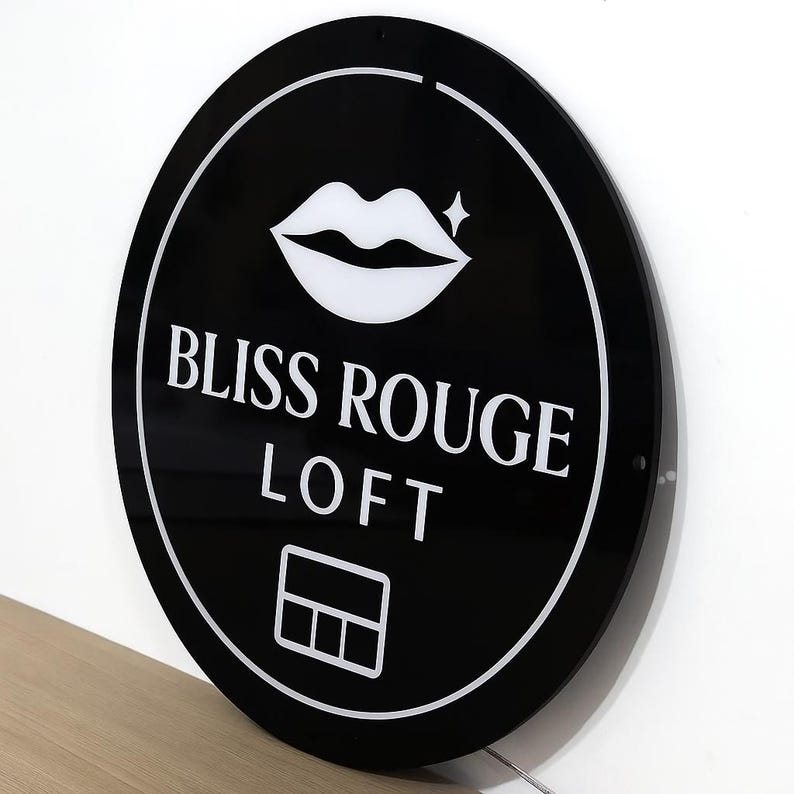 May include: A round, black sign with white text and a white outline. The sign reads "BLISS ROUGE LOFT" with a graphic of lips and a small star. A white graphic of a card is at the bottom of the sign.