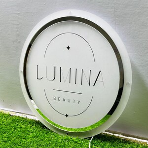 K&ouml;nnte beinhalten: Ein rundes, beleuchtetes Schild mit dem Wort "LUMINA" in Schwarz, &uuml;ber "BEAUTY". Das Schild hat eine wei&szlig;e Oberfl&auml;che, einen silberfarbenen Rand und einen wei&szlig;en Au&szlig;enring. Das Schild befindet sich auf einer gr&uuml;nen Oberfl&auml;che.