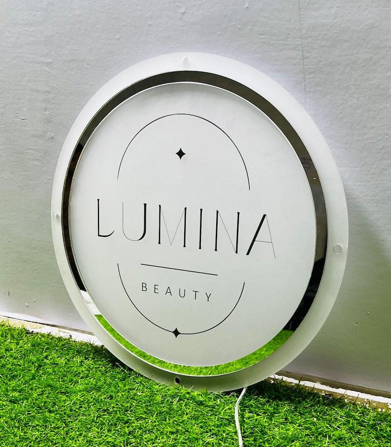 K&ouml;nnte beinhalten: Ein rundes, beleuchtetes Schild mit dem Wort "LUMINA" in Schwarz, &uuml;ber "BEAUTY". Das Schild hat eine wei&szlig;e Oberfl&auml;che, einen silberfarbenen Rand und einen wei&szlig;en Au&szlig;enring. Das Schild befindet sich auf einer gr&uuml;nen Oberfl&auml;che.