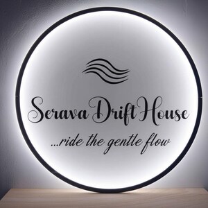 May include: A round, illuminated sign with a black border. The sign features the words "Serava Drift House" in elegant script, along with the phrase "...ride the gentle flow." A stylized wave graphic is above the text.