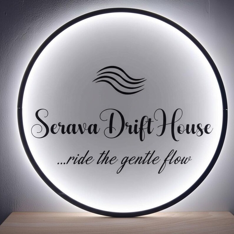 May include: A round, illuminated sign with a black border. The sign features the words "Serava Drift House" in elegant script, along with the phrase "...ride the gentle flow." A stylized wave graphic is above the text.