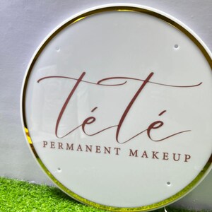 K&ouml;nnte beinhalten: Rundes, wei&szlig;es Schild mit goldenem Rand und dem burgunderfarbenen Text "T&eacute;t&eacute; PERMANENT MAKEUP". Das Schild ist beleuchtet und liegt auf gr&uuml;nem Kunstrasen.