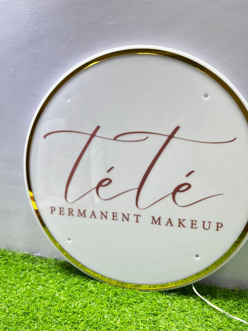 K&ouml;nnte beinhalten: Rundes, wei&szlig;es Schild mit goldenem Rand und dem burgunderfarbenen Text "T&eacute;t&eacute; PERMANENT MAKEUP". Das Schild ist beleuchtet und liegt auf gr&uuml;nem Kunstrasen.