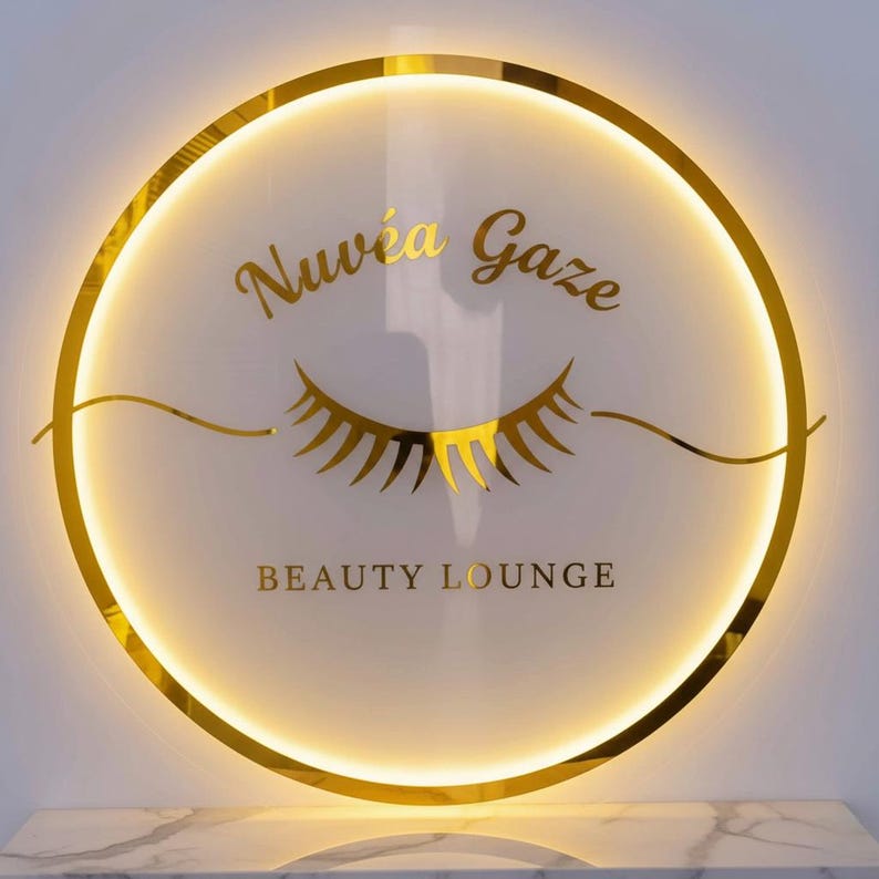May include: A round, illuminated sign with a gold border and the words "Nuv&eacute;a Gaze BEAUTY LOUNGE" in gold lettering. The sign features a gold eyelash design. The background is white, and the sign is mounted on a marble surface.
