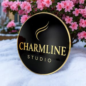 May include: A round, black sign with gold lettering and a gold border. The sign reads "CHARMLINE STUDIO" in a serif font. The background features pink flowers and snow.