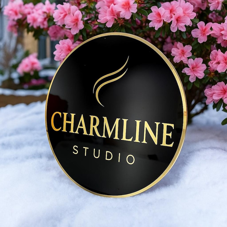 May include: A round, black sign with gold lettering and a gold border. The sign reads "CHARMLINE STUDIO" in a serif font. The background features pink flowers and snow.