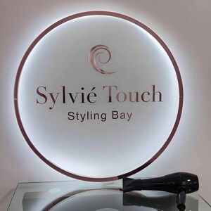 May include: A round, illuminated sign with a rose gold border and the text "Sylvie Touch Styling Bay." A black hair dryer rests on a glass surface below the sign, suggesting a hair salon or styling business.