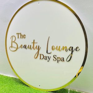 May include: A circular white sign with a gold-coloured border. The words "The Beauty Lounge Day Spa" are in gold script across the centre. The sign is set against a green and white background.