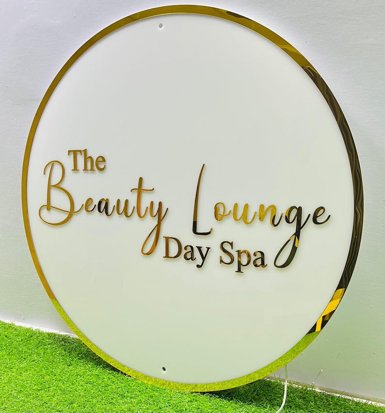 May include: A circular white sign with a gold-coloured border. The words "The Beauty Lounge Day Spa" are in gold script across the centre. The sign is set against a green and white background.