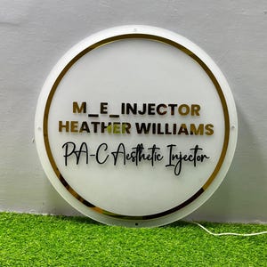 K&ouml;nnte beinhalten: Ein rundes, wei&szlig;es Schild mit goldenem Rand und Schriftzug. Der Text lautet "M_E_INJECTOR HEATHER WILLIAMS PA-C Aesthetic Injector" in Gold und Schwarz. Das Schild wird vor einem gr&uuml;nen Hintergrund gezeigt.