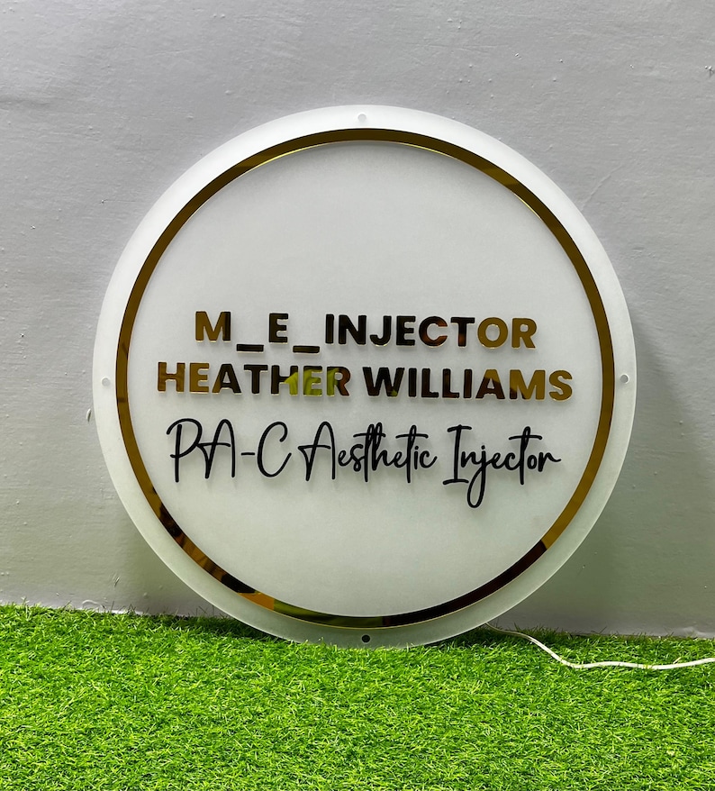 K&ouml;nnte beinhalten: Ein rundes, wei&szlig;es Schild mit goldenem Rand und Schriftzug. Der Text lautet "M_E_INJECTOR HEATHER WILLIAMS PA-C Aesthetic Injector" in Gold und Schwarz. Das Schild wird vor einem gr&uuml;nen Hintergrund gezeigt.