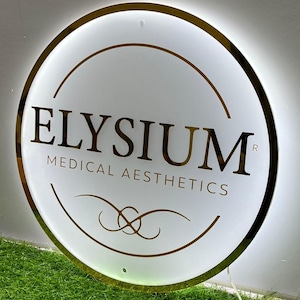 May include: A circular, illuminated sign with a gold trim and the words "ELYSIUM MEDICAL AESTHETICS" in gold. The sign is white with gold design accents and is lit from behind. The sign is on a green surface.