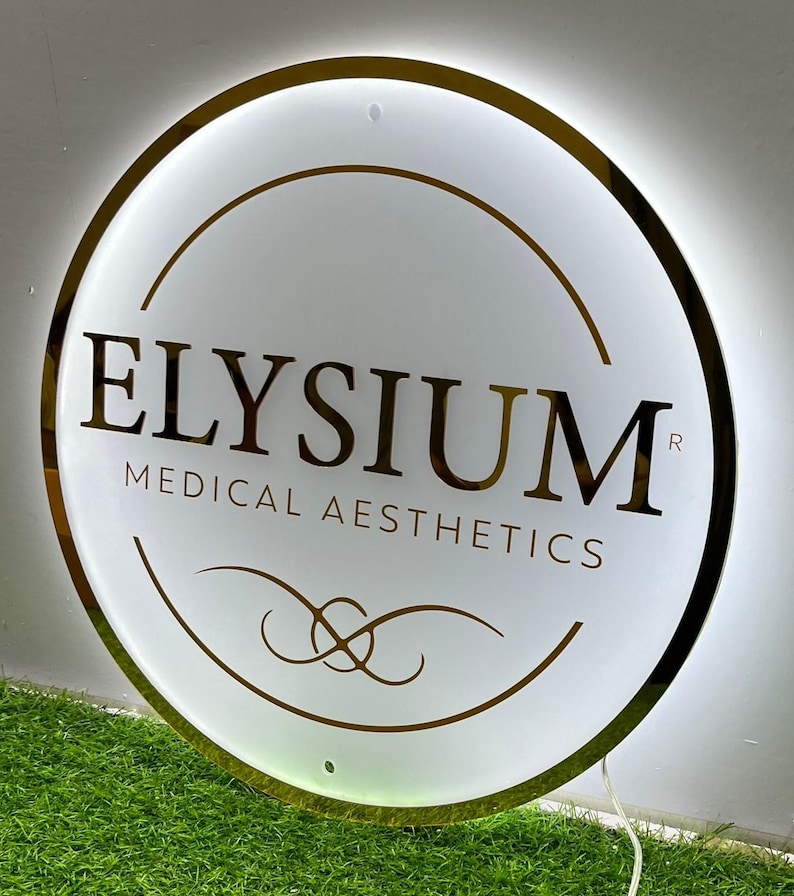 May include: A circular, illuminated sign with a gold trim and the words "ELYSIUM MEDICAL AESTHETICS" in gold. The sign is white with gold design accents and is lit from behind. The sign is on a green surface.