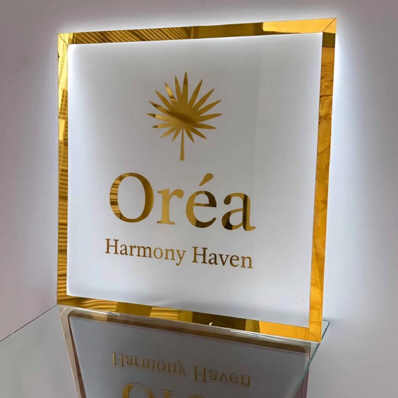 May include: A square sign with a gold frame and a white interior. The sign features the gold text "Orea" above "Harmony Haven" and a gold palm leaf design. The sign is reflected on a glass surface.