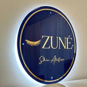 May include: A round, navy blue sign with gold trim and backlighting. The sign features the name "ZUN&Eacute;" in gold lettering, along with the words "Skin Atelier" and a gold eyelash design. The sign is mounted on a white wall.
