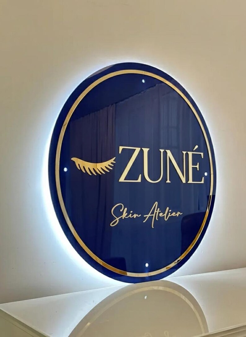 May include: A round, navy blue sign with gold trim and backlighting. The sign features the name "ZUN&Eacute;" in gold lettering, along with the words "Skin Atelier" and a gold eyelash design. The sign is mounted on a white wall.