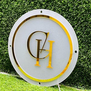 K&ouml;nnte beinhalten: Ein runder, wei&szlig;er Leuchtschild mit goldenem Rand und goldenen Buchstaben. Das Schild zeigt die goldenen Buchstaben "CH" in einer stilisierten Schriftart. Das Schild befindet sich vor einem Hintergrund aus gr&uuml;nem Gras.