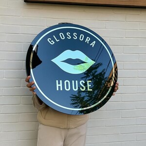 May include: A round, black sign with the words "GLOSSORA HOUSE" in white lettering. The sign features a stylized, light blue lip graphic. The sign is held in front of a white brick wall.