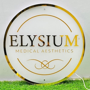 May include: A circular illuminated sign with a gold frame and lettering. The sign displays the word "ELYSIUM" in gold and black, with "MEDICAL AESTHETICS" underneath. Decorative gold lines and a gold border complete the design.