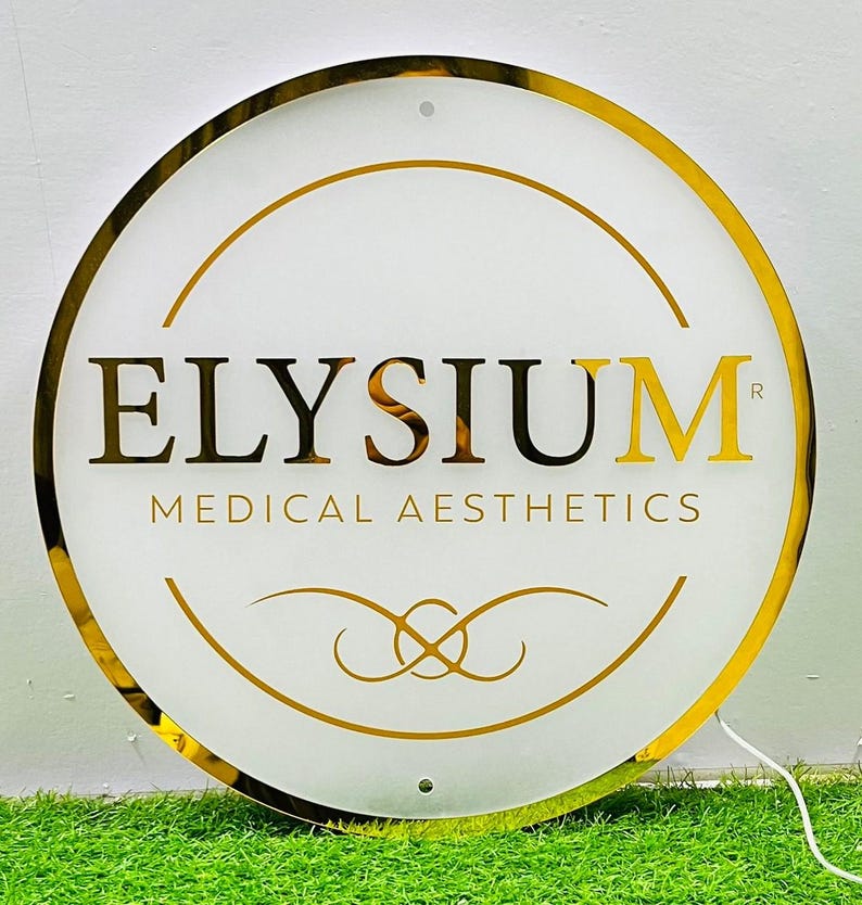 May include: A circular illuminated sign with a gold frame and lettering. The sign displays the word "ELYSIUM" in gold and black, with "MEDICAL AESTHETICS" underneath. Decorative gold lines and a gold border complete the design.