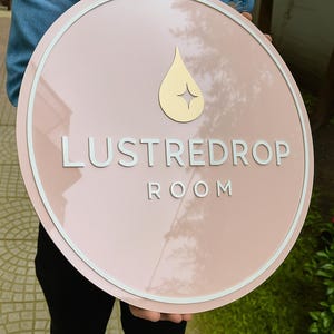 May include: A round, pink sign with the text "LUSTREDROP ROOM" in white, and a gold teardrop design. The sign has a white border and is held by a person wearing a denim jacket.