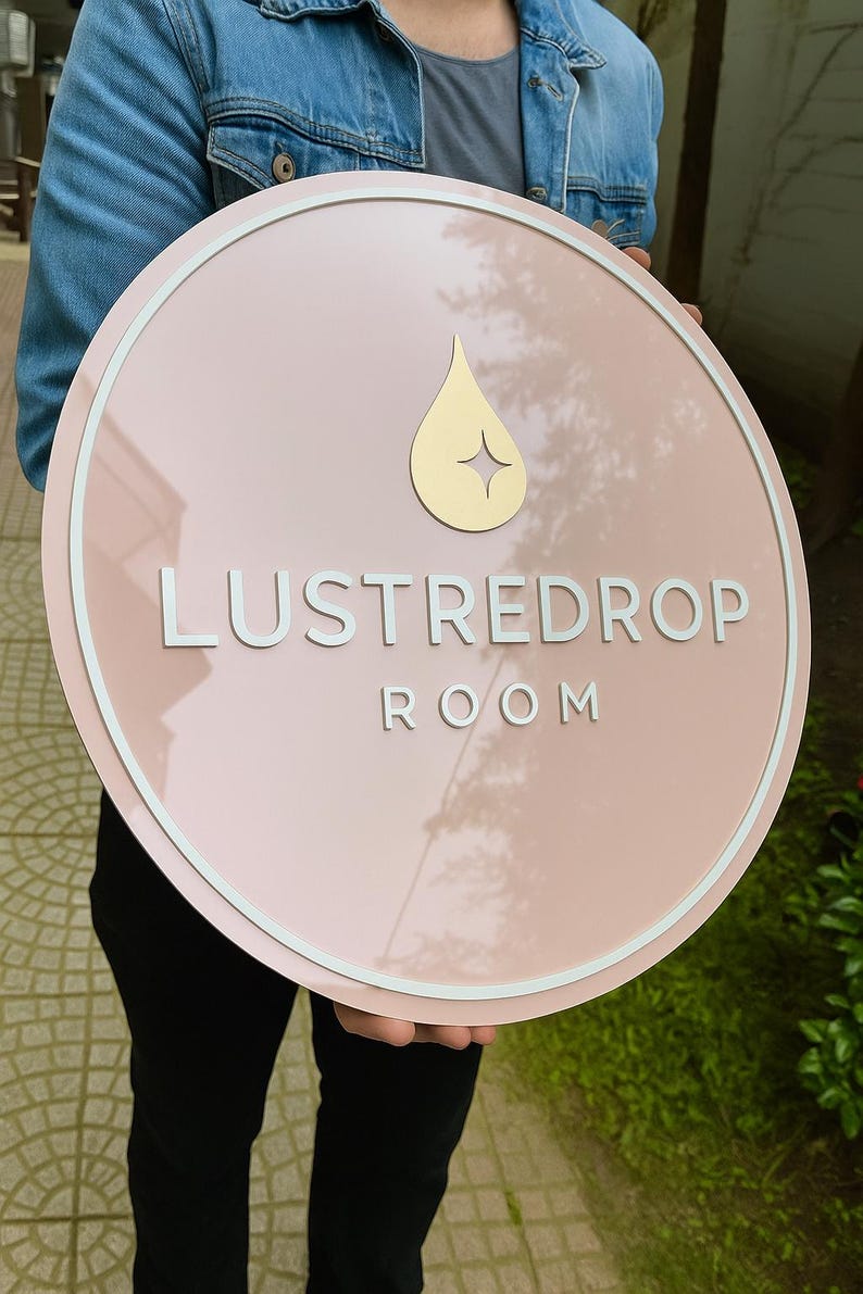 May include: A round, pink sign with the text "LUSTREDROP ROOM" in white, and a gold teardrop design. The sign has a white border and is held by a person wearing a denim jacket.