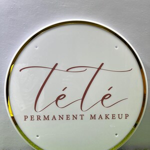 K&ouml;nnte beinhalten: Rundes, wei&szlig;es Schild mit goldenem Rand. Der Schriftzug "T&eacute;t&eacute;" in bordeauxfarbener Schrift ist zu sehen, darunter "PERMANENT MAKEUP" in einer Serifenschrift. Das Schild ist beleuchtet und hat eine gl&auml;nzende Oberfl&auml;che.