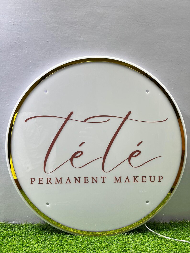 K&ouml;nnte beinhalten: Rundes, wei&szlig;es Schild mit goldenem Rand. Der Schriftzug "T&eacute;t&eacute;" in bordeauxfarbener Schrift ist zu sehen, darunter "PERMANENT MAKEUP" in einer Serifenschrift. Das Schild ist beleuchtet und hat eine gl&auml;nzende Oberfl&auml;che.